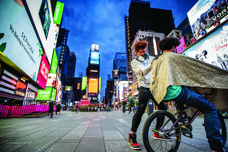 Julien Howard cutting the hair of a man sitting on a BMX bicycle at night in Times Square while wearing a face mask.