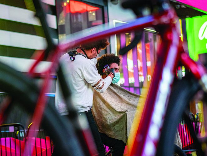 A Trek bicycle in the foreground in front of Julien Howard cutting the hair of a client with city billboards behind them.