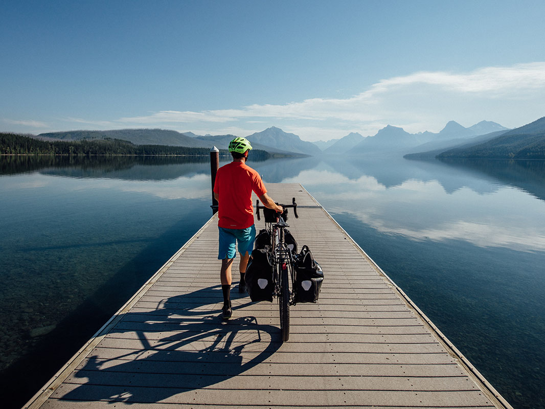 Adventure cyclist walks bike with panniers on a dock going into a lake with mountain range in distance.