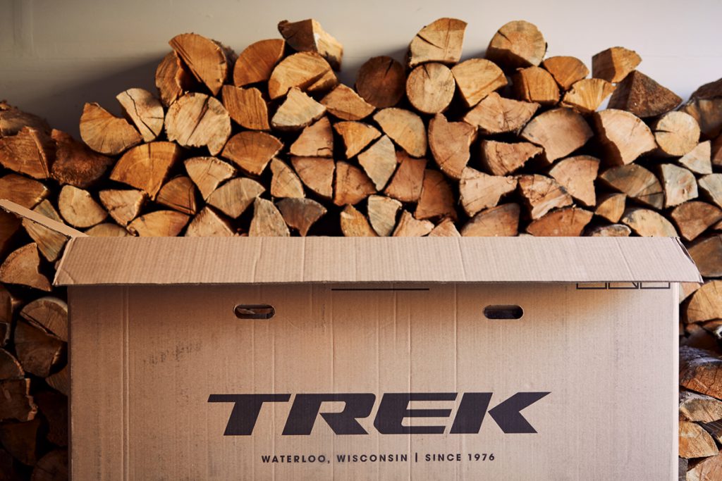 A cardboard Trek bike box in front of a stack of chopped wood.