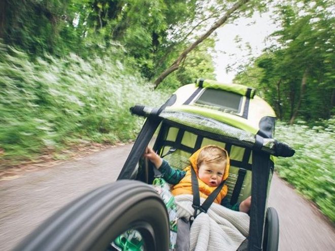 A child wearing a yellow vest rides behind a bicycle in a trailer on a dirt trail in a forest.