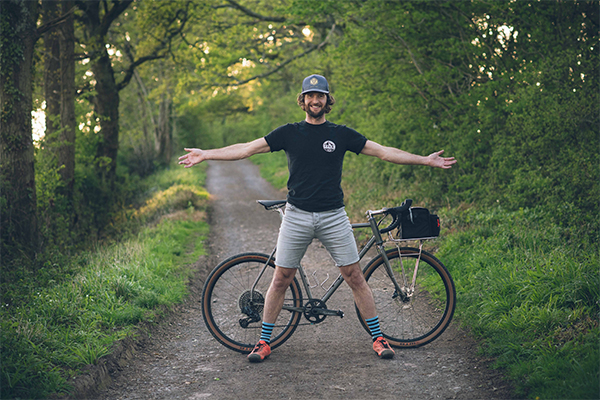 Wayne Reif posing on a dirt track in the woods. He is standing in front of his bicycle with his arms spread open.