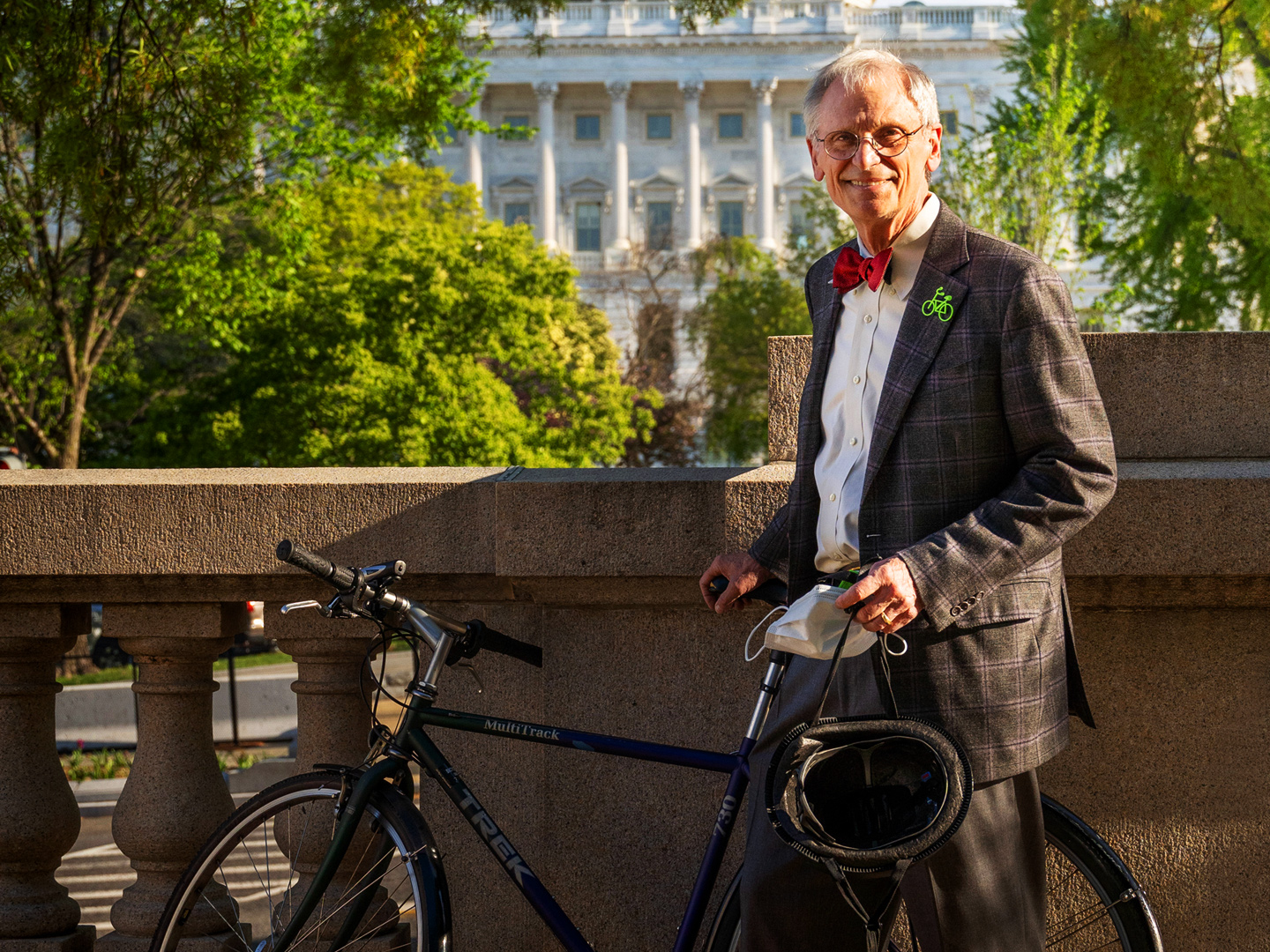 Earl Blumenauer stands holding his bicycle. In the background are trees and the White House.