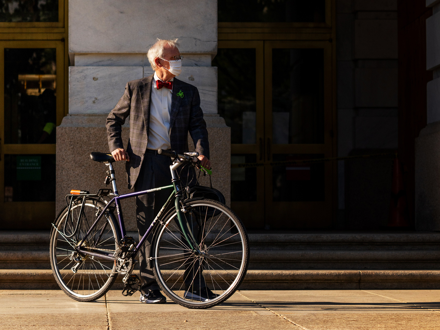 Congressman Earl Blumenauer stands behind his green bicycle. He is wearing a grey suit with a red bowtie and a white surgical mask. He is holding his green bicycle in front of him, and stands on a sidewalk with a grey building behind him.