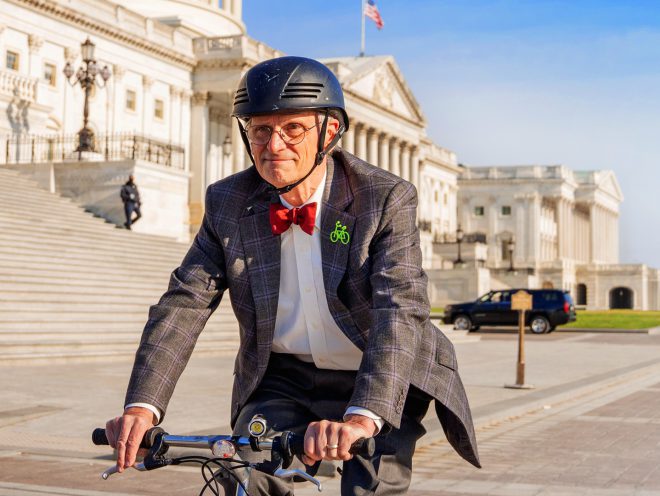 Congressman Earl Blumenauer rides his bike in front of the US Capitol building. He is wearing a bicycle helmet and suit with a bright red bowtie.