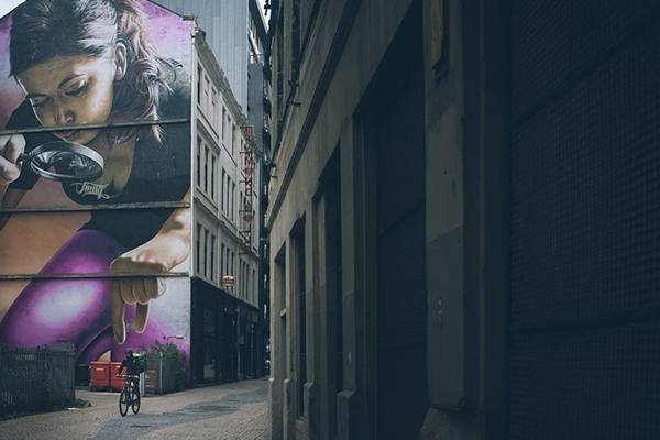 A photo of a cyclist riding down a dim back alleyway. Behind the cyclist is a large wall mural of a girl with a magnifying glass picking up something between her finger and thumb. The photo is taken so that it looks like the girl is about to pick up the cyclist in the foreground.