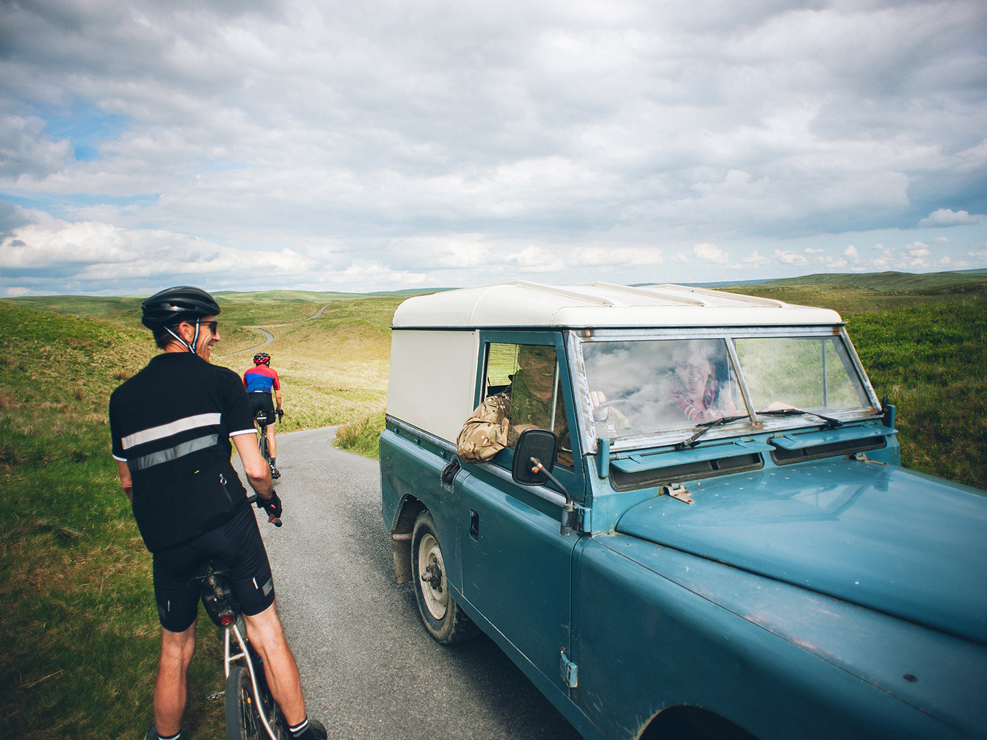 Pyöräilijä, joka on pysähtynyt vintage-jeepin viereen juttelemaan ajajan kanssa