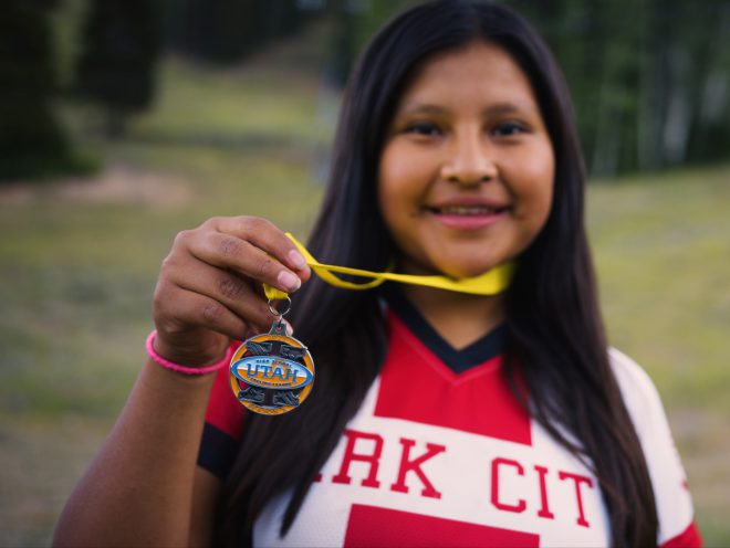 Genesis is smiling and showing her gold medal to the camera.