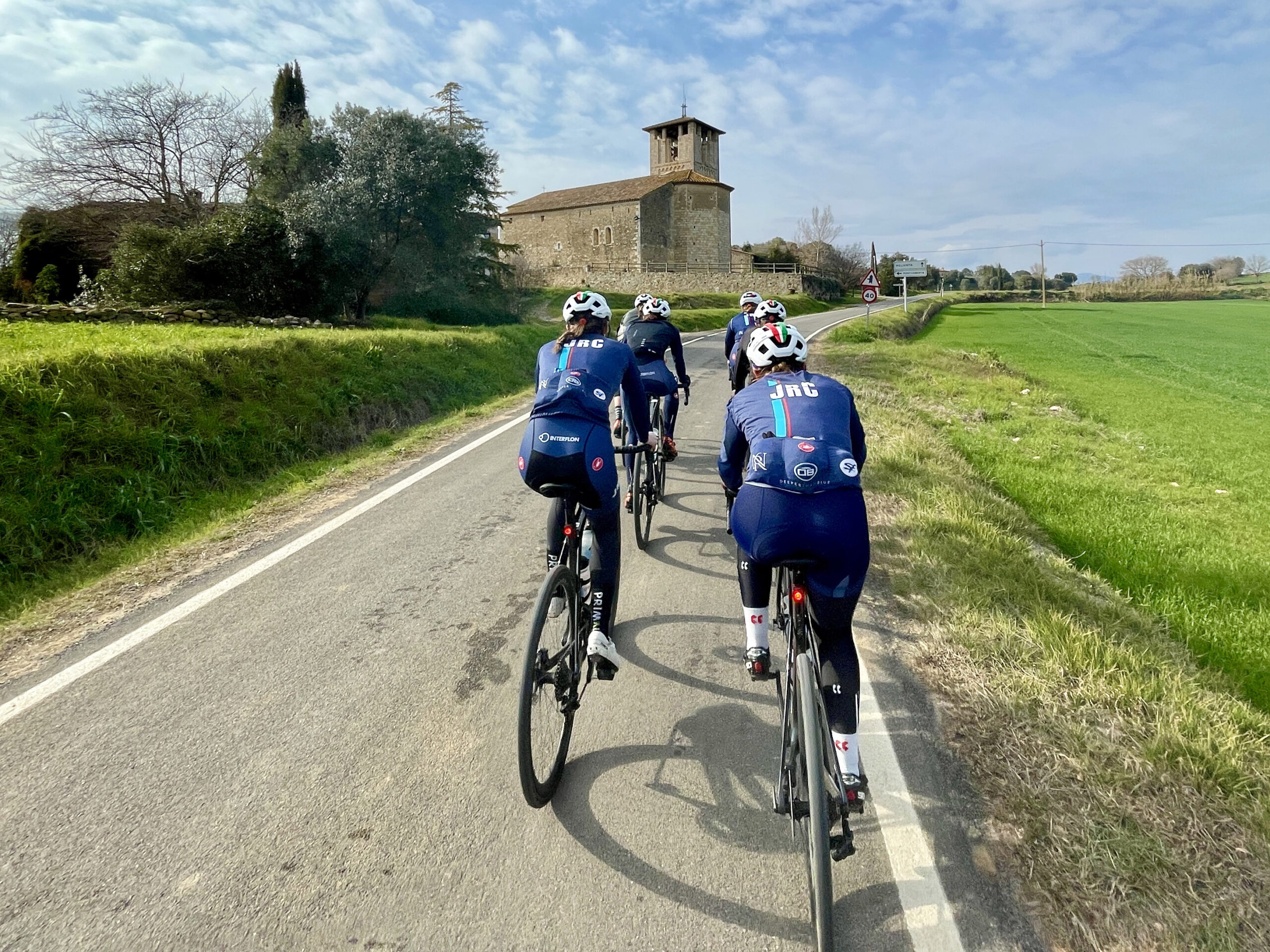 Riders from JRC Women's Junior Cycling team riding away from the camera on a paved road toward an old stone building in Girona.
