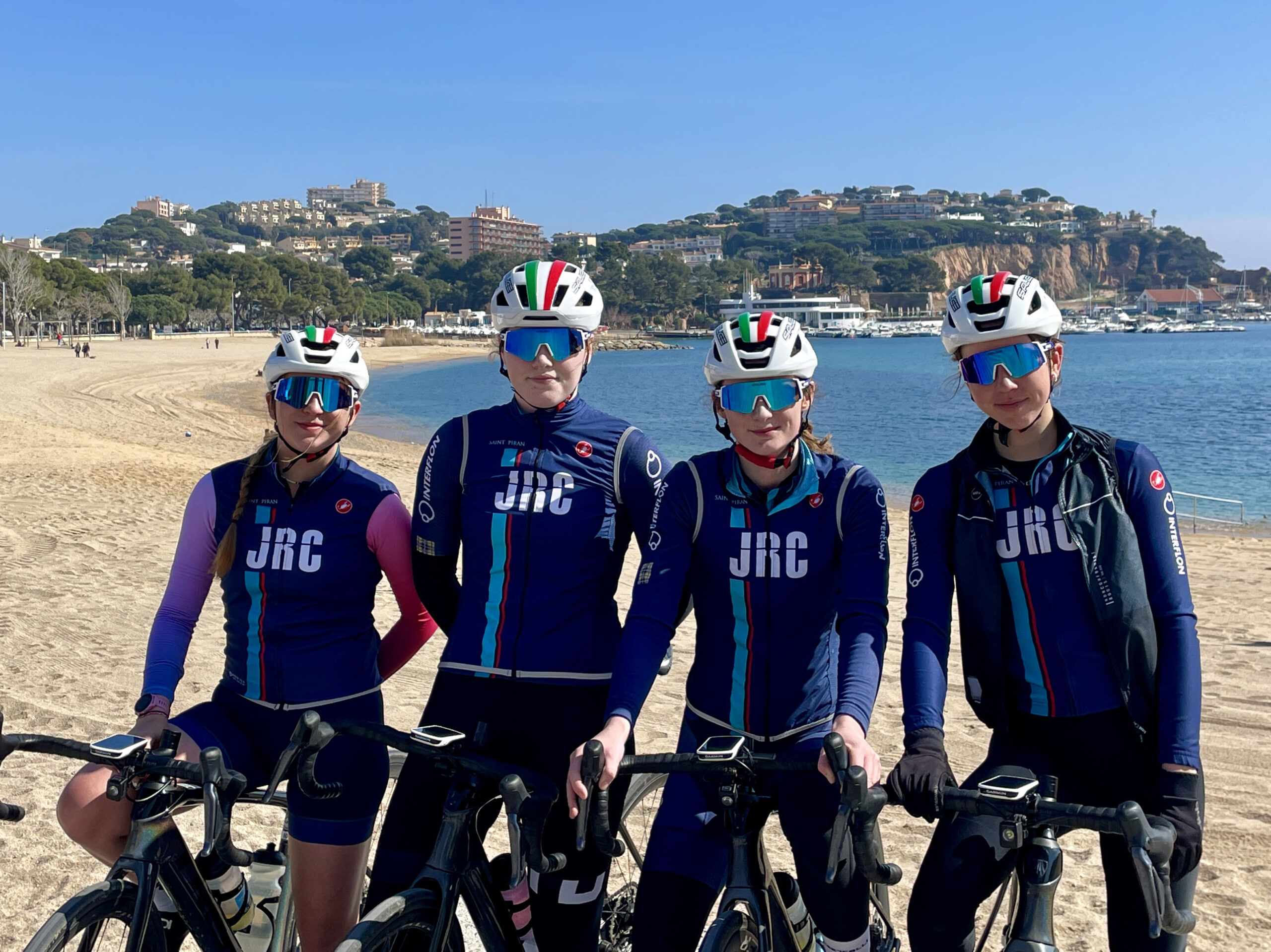 Four riders from the JRC Women's Junior Cycling team standing over their bikes in front of a Spanish coastline.