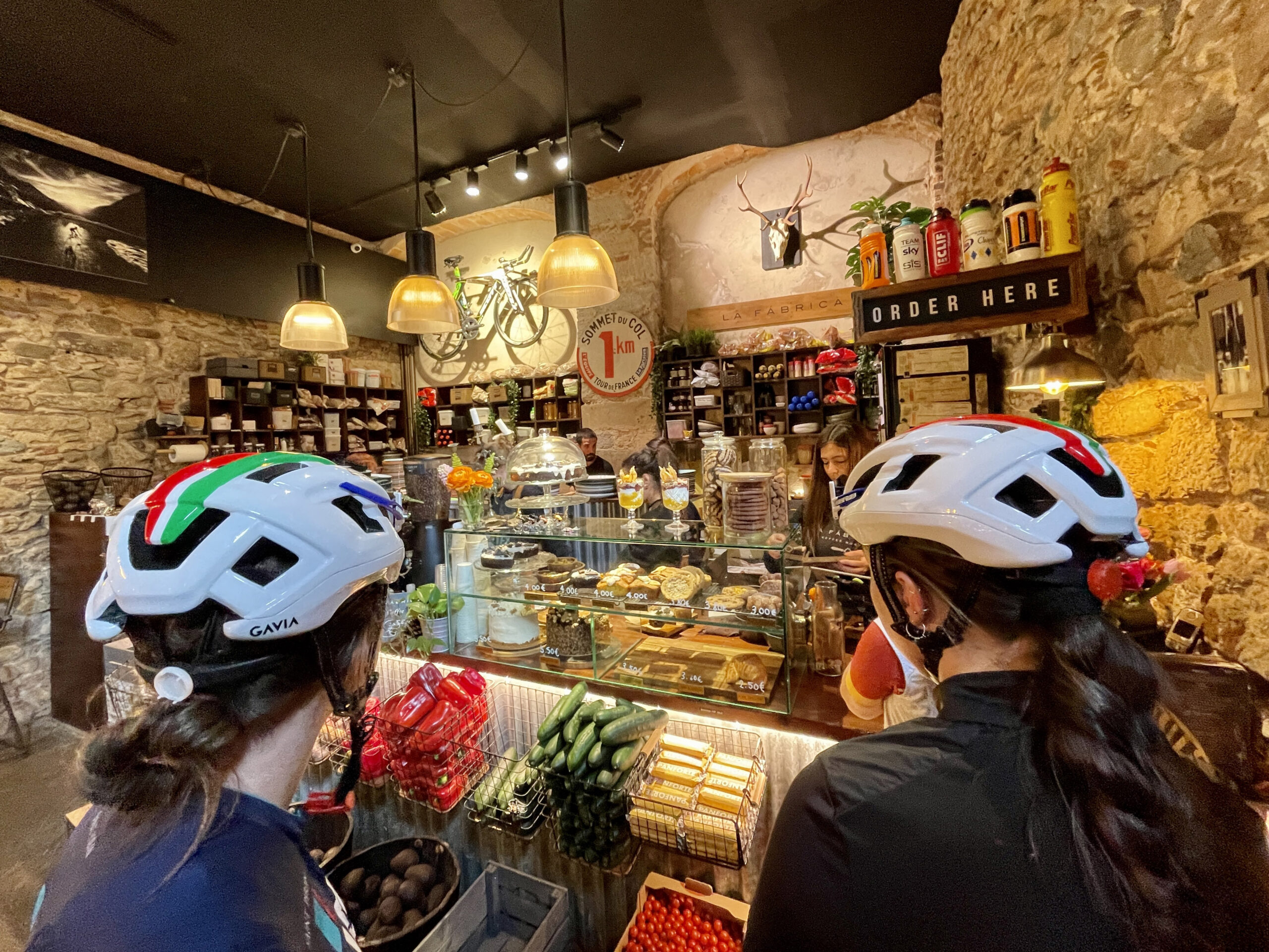 Two women in helmets look at a case of desserts.