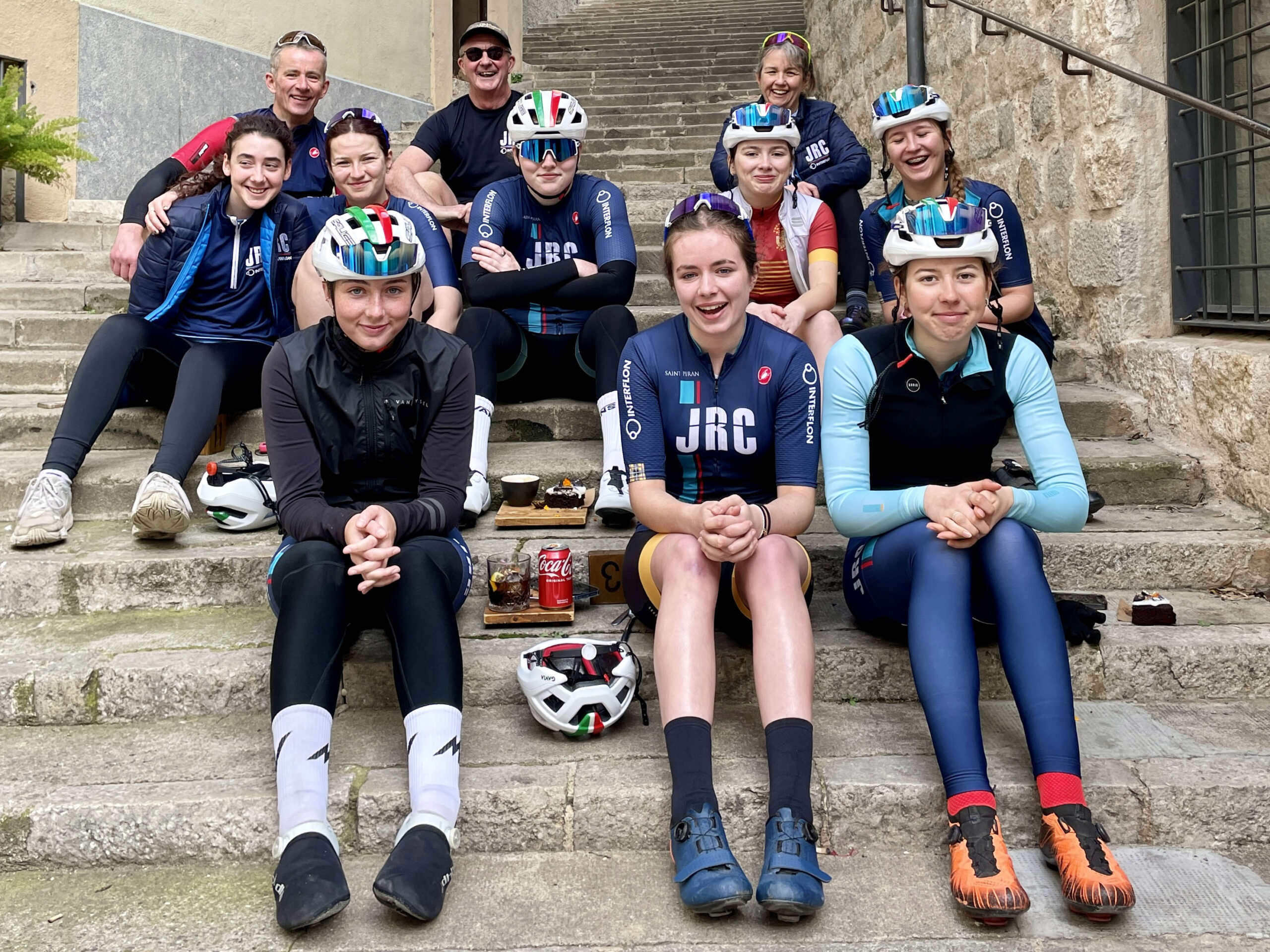 The JRC Women's Junior Cycling team wearing their team kits and sitting on concrete steps in Girona.