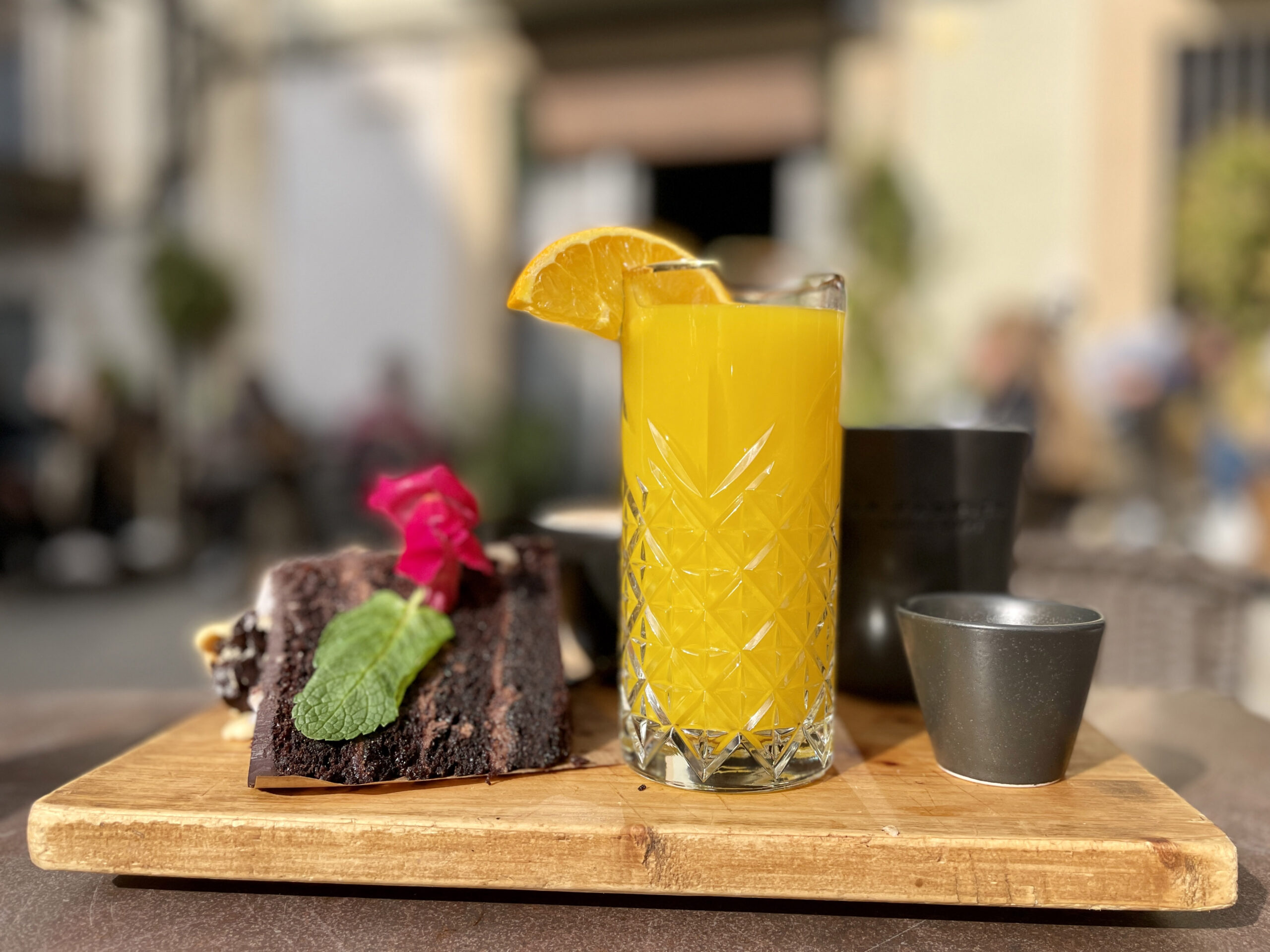 A piece of chocolate cake on a wood board next to a fancy orange drink.