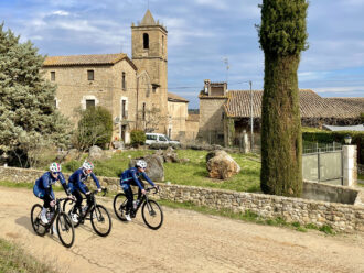Three women riding road bikes on a dirt road in front of an old stone building in Girona.
