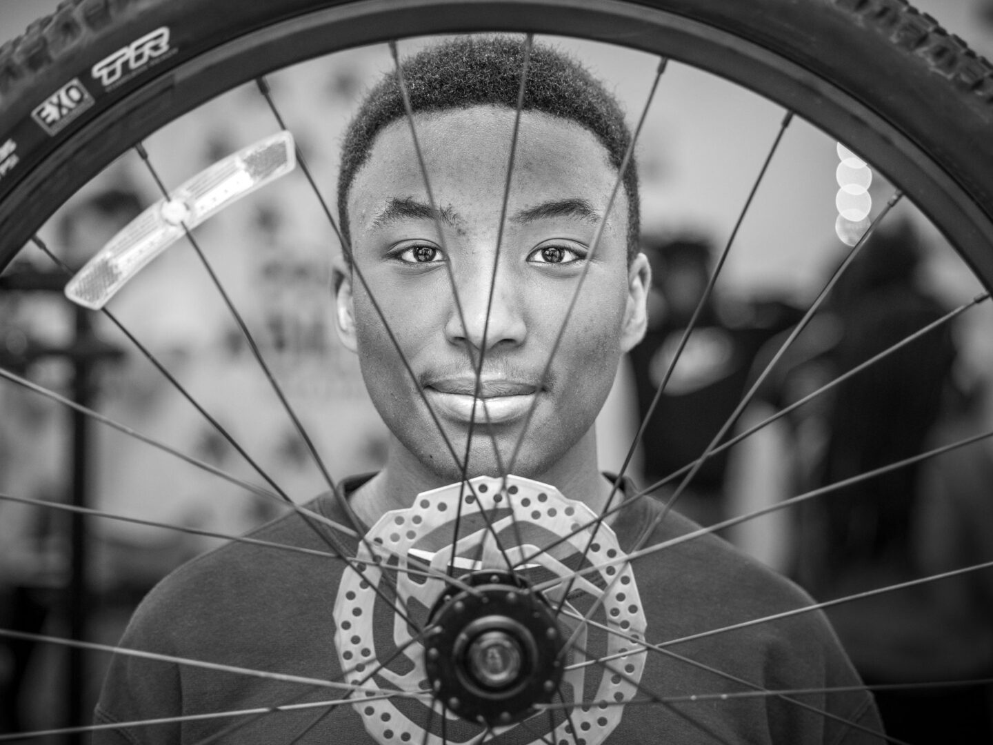 A boy's smiling face behind the spokes of a bicycle wheel.