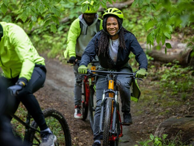 A smiling boy riding his mountain bike on a dirt path with green trees and another rider behind him.