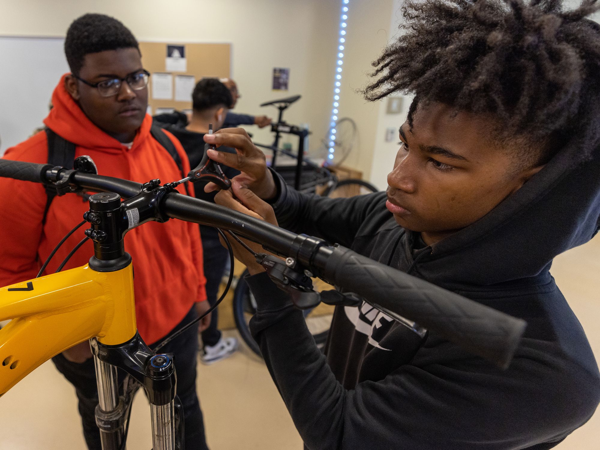A student looks on as another works on the handlebar of a bike.