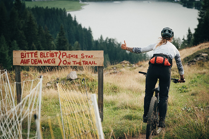 Mountainbikerin auf einem Trail, daneben ein Holzschild mit der Aufschrift 
